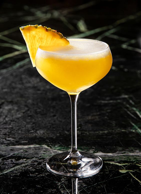 cocktail aguardente de medronho com amarguinha e sumo de ananas. decoração no topod to copo com triangulo de ananas. fundo marmore escuro. copo alto com liquido amarelo alaranjado